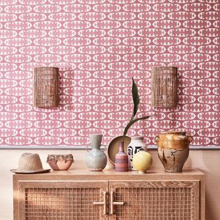 A hallway with the top half of a wall covered in a red-toned printed wallpaper with a console table displaying decorative objects in front of it