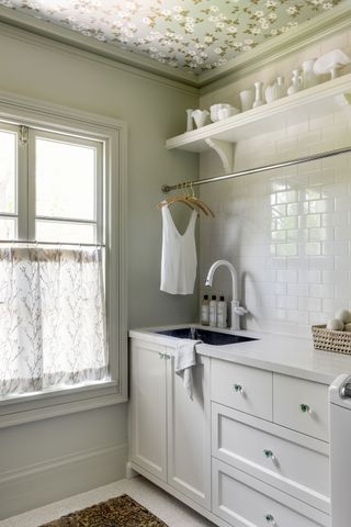 softly lit laundry space with fitted cupboards and lace curtain with hanging rail and tiled splashback and floral wallpaper on the ceiling