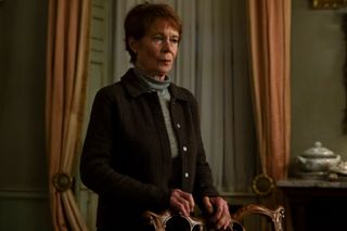 Celia Imrie as Margaret Roylin, resting her hands on the back of a chair while standing in a parlor, in episode 204 of The Diplomat.