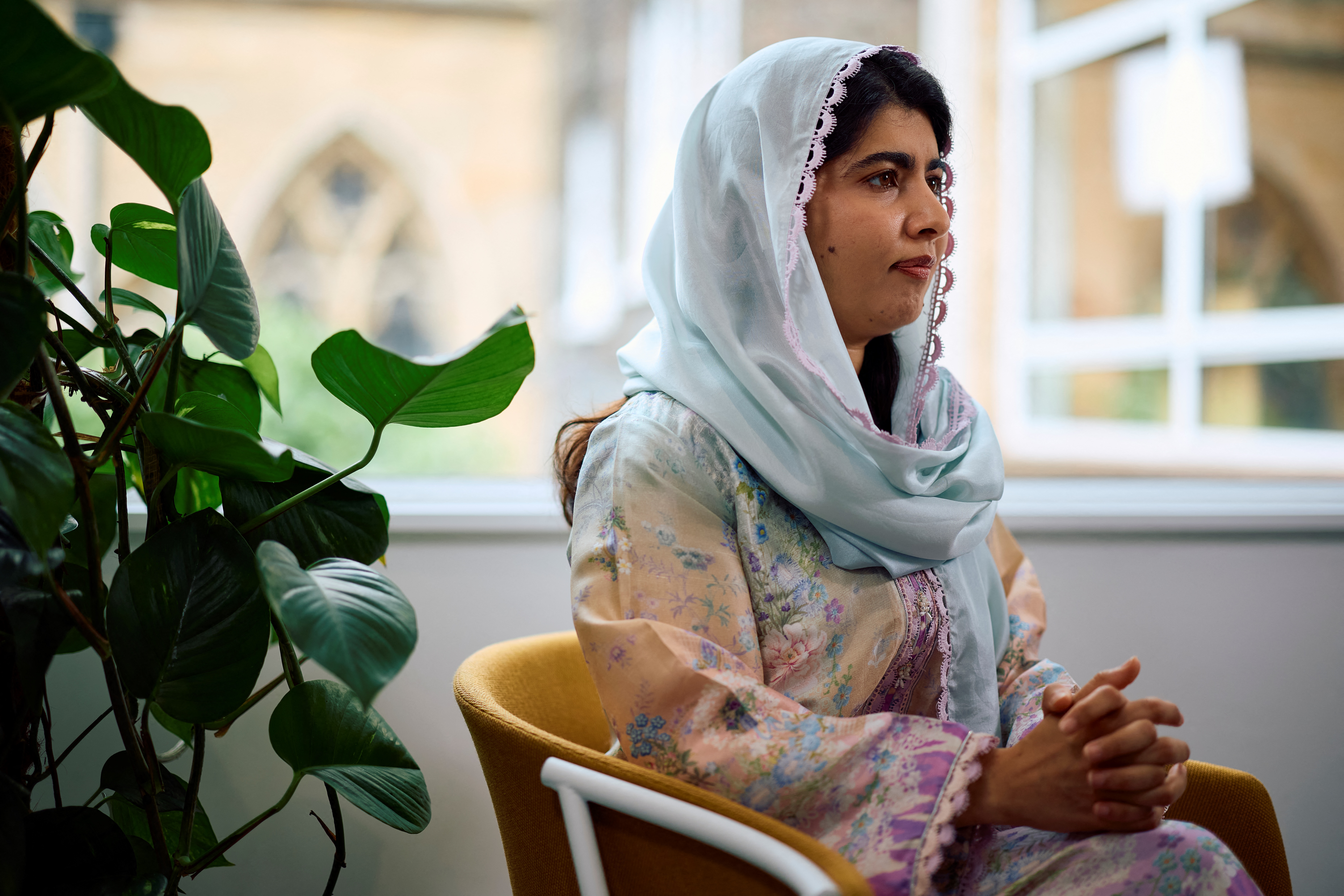 Malala Yousafzai sits near a window in a light blue headscarf and floral dress during an interview, hands clasped in her lap.