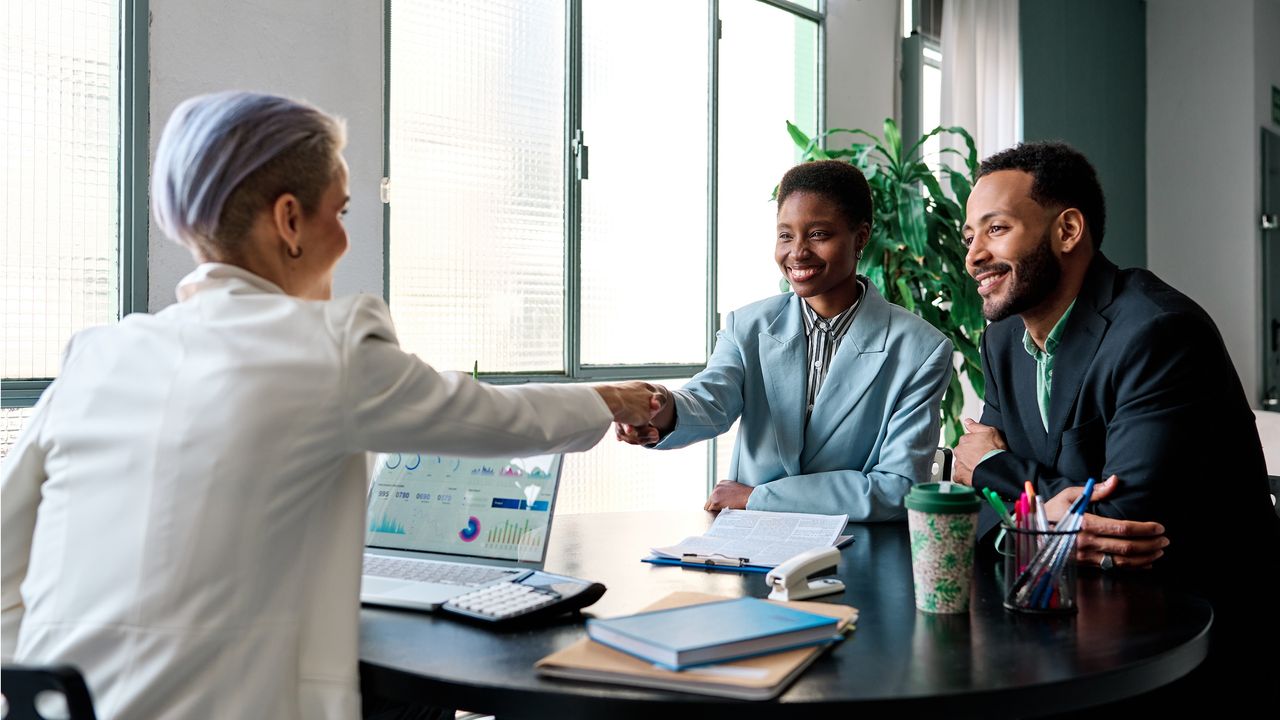 A financial adviser shakes hands across her desk with a client couple.