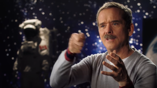 chris hadfield talking. to his left and behind him is a spacesuit with a canadian flag sewn on the arm