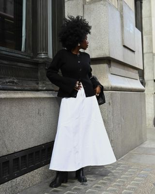 fashion influencer Danielle Oreoluwa Jinadu poses on the street wearing a black sweater-jacket, full white maxi skirt, and ankle boots