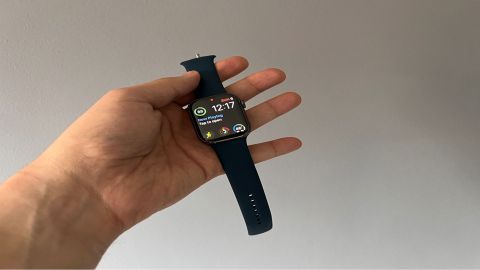 Image shows a hand holding the Apple Watch Series 7 with a blue strap. The watchface is turned on.