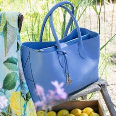 A blue Aspinal of London hang bag on a wooden stool in a sunny outdoor setting.