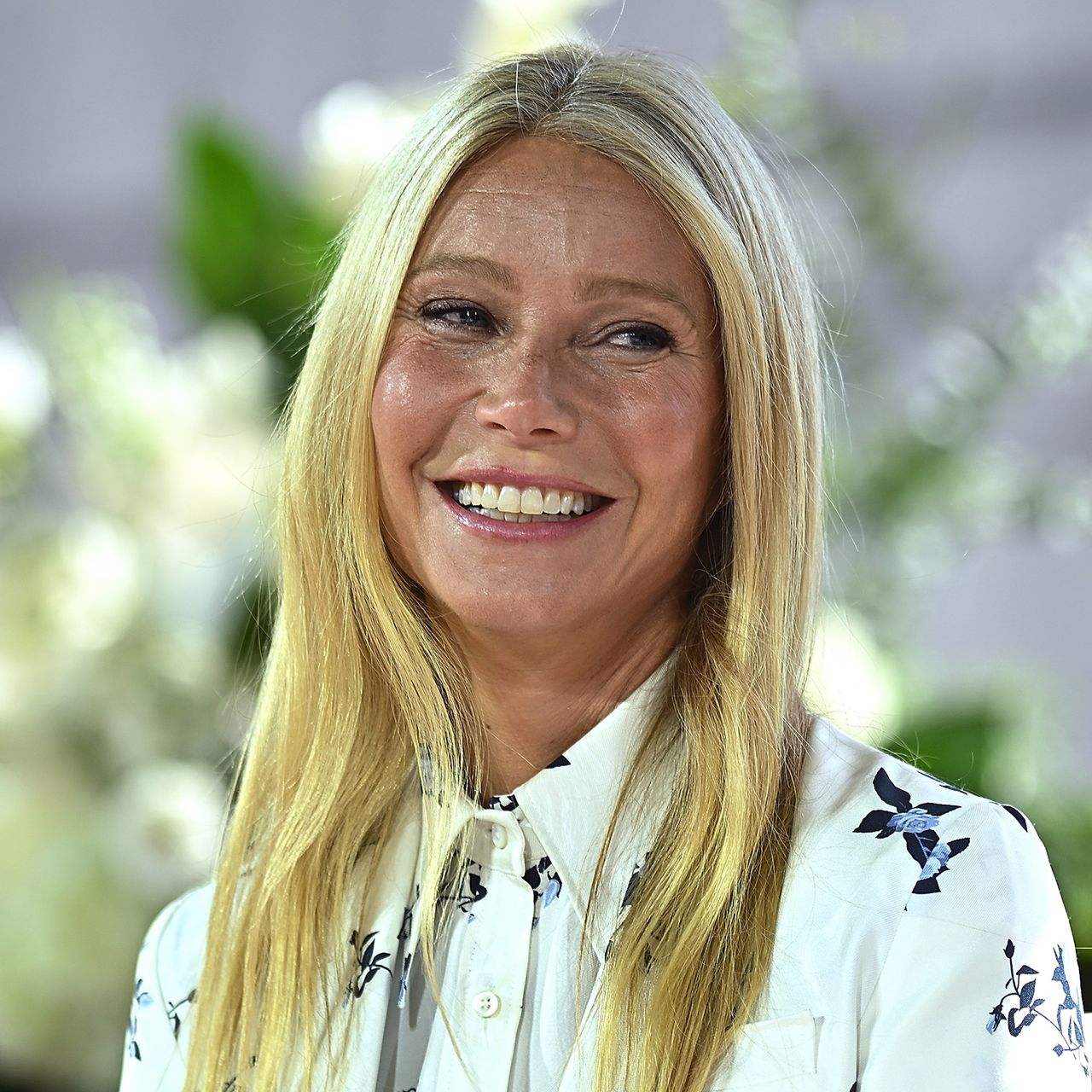 Headshot of Gwyneth Paltrow speaking at Forbes Power Women&#039;s Summit, wearing white blouse with foliage visible in background