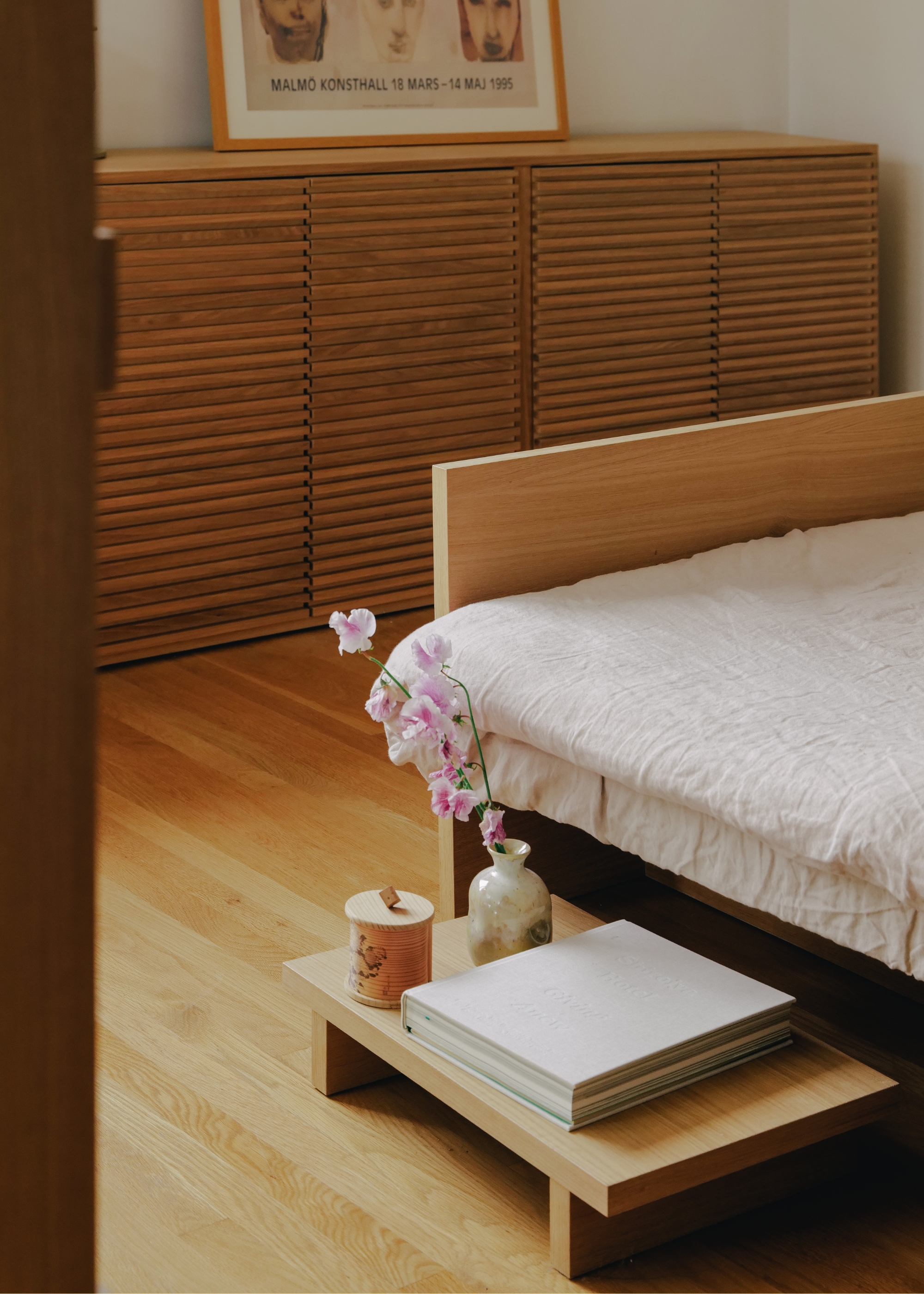 A wood bed with a low mini desk with a vase of flowers, a book, and a candle