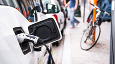 A white electric car is being charged, parked. In the background, a bicycle is resting against an orange pole.