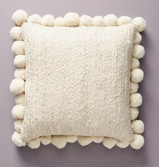 A cream cushion with a pom pom trim on a lilac background.