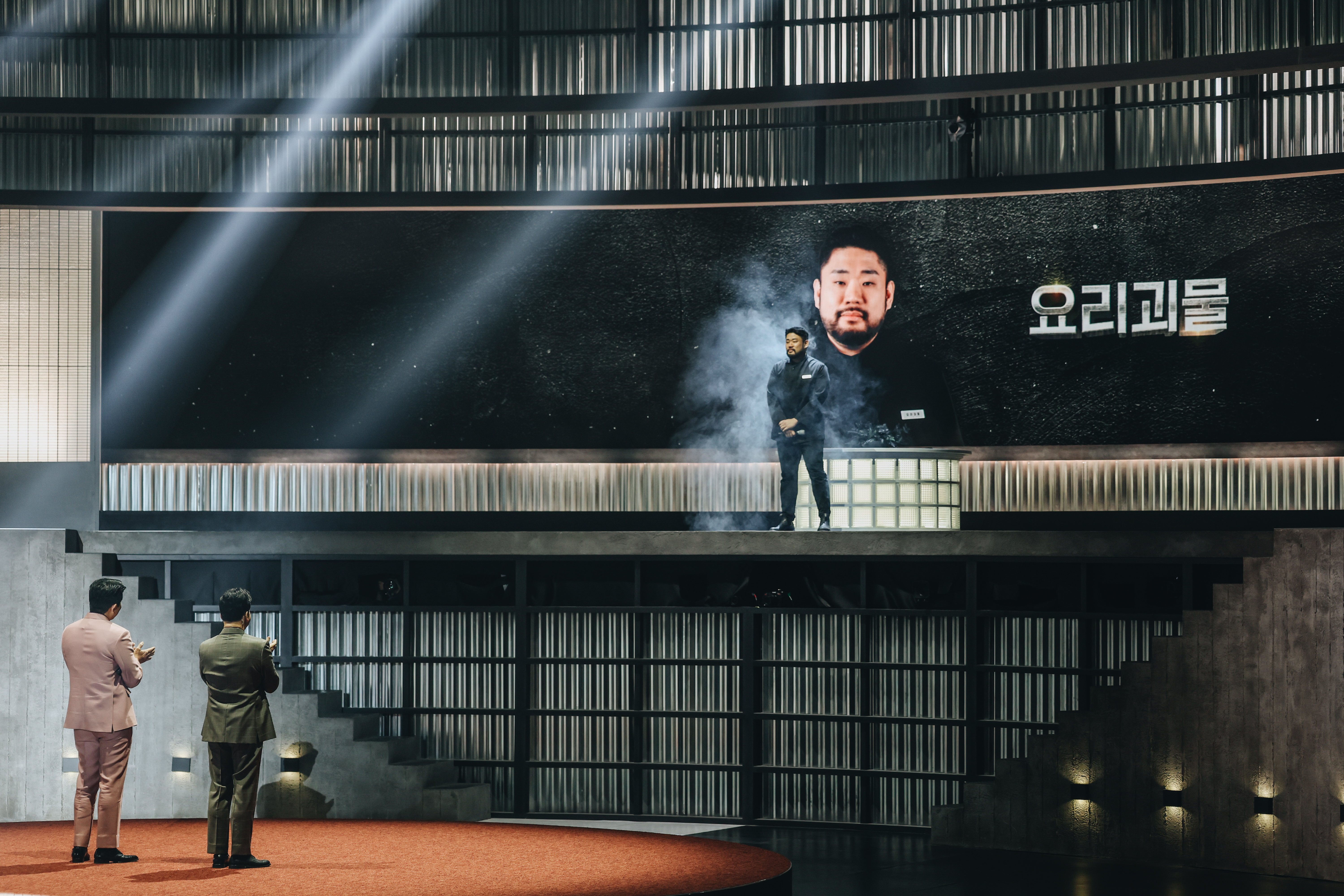 A man (Hasung Lee) stands on an elevated stage as two men applaud, in the 'Culinary Class Wars' season 2 finale.