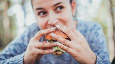 Woman is shown eating a burger. She is looking into the distance. The background is blurry.