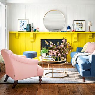 a living room with two tone wall with white on top, separated by a full length yellow shelf with yellow wall underneath, with a pink armchair, blue couch and gold nested coffee tables