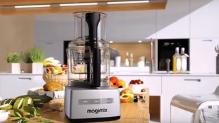 A Magimix food processor on a kitchen countertop surrounded by fresh produce