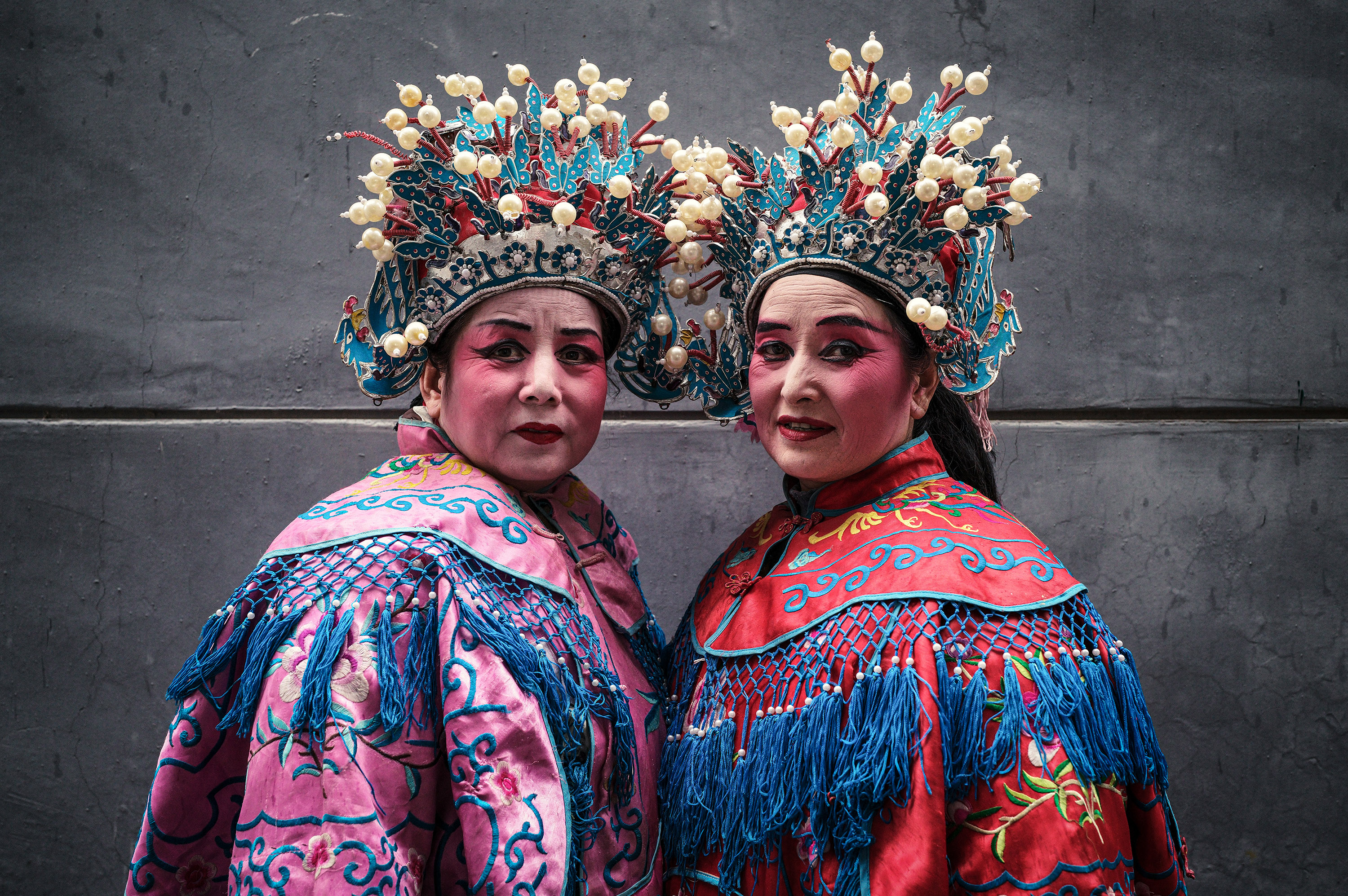 Two performers in vibrant pink and red robes with elaborate blue and gold headdresses studded with pearls turn to face the camera against a grey concrete wall.