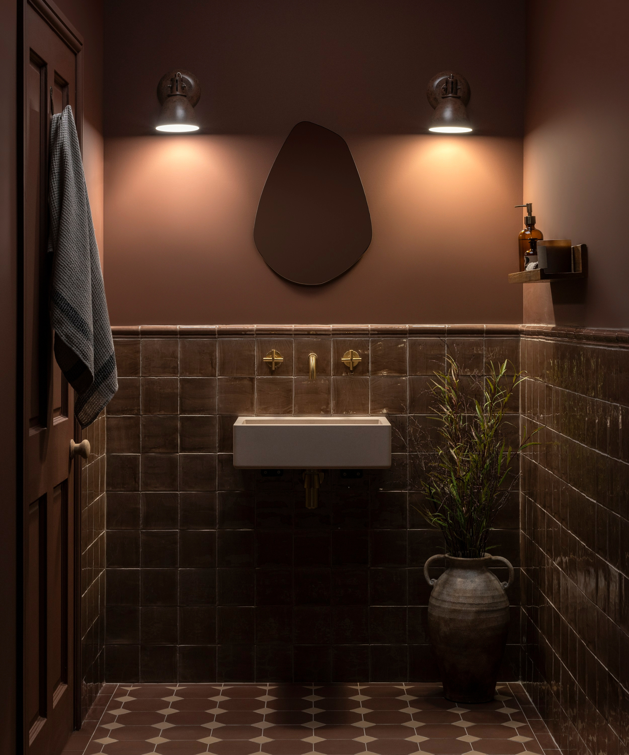 A moody dark bathroom with deep brown glazed square wall tiles, a geometric patterned floor, wall-mounted white basin with brass taps, organic-shaped mirror and twin industrial wall lights.