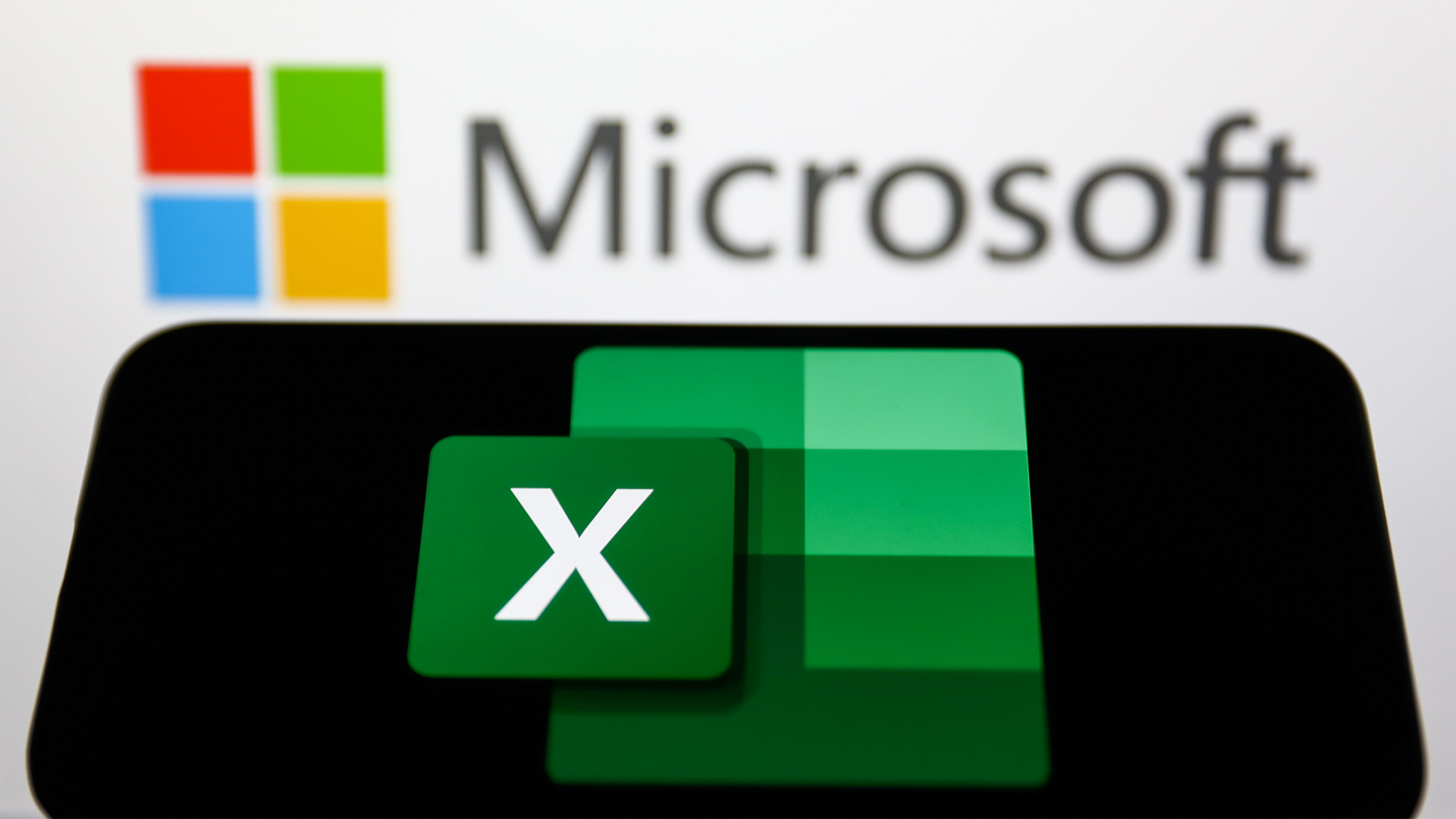 Microsoft Excel logo pictured on a smartphone screen in foreground with Microsoft branding in background.