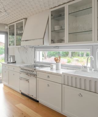 things that make a kitchen look cheap, all white kitchen with chrome fixtures and fittings, textured ceiling, cooker hood, marble countertops, Butler sink, glazed wall units, mid toned wooden floor