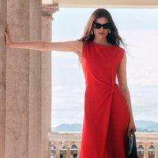 A woman posing in a red dress with black sunglasses and a black leather handbag leaning with one arm against a Roman-style pillar in front of a mountainous view