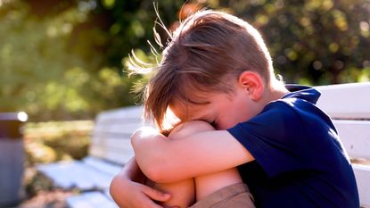 Sad little boy sitting on a bench outdoors clutching his knees and burying his face