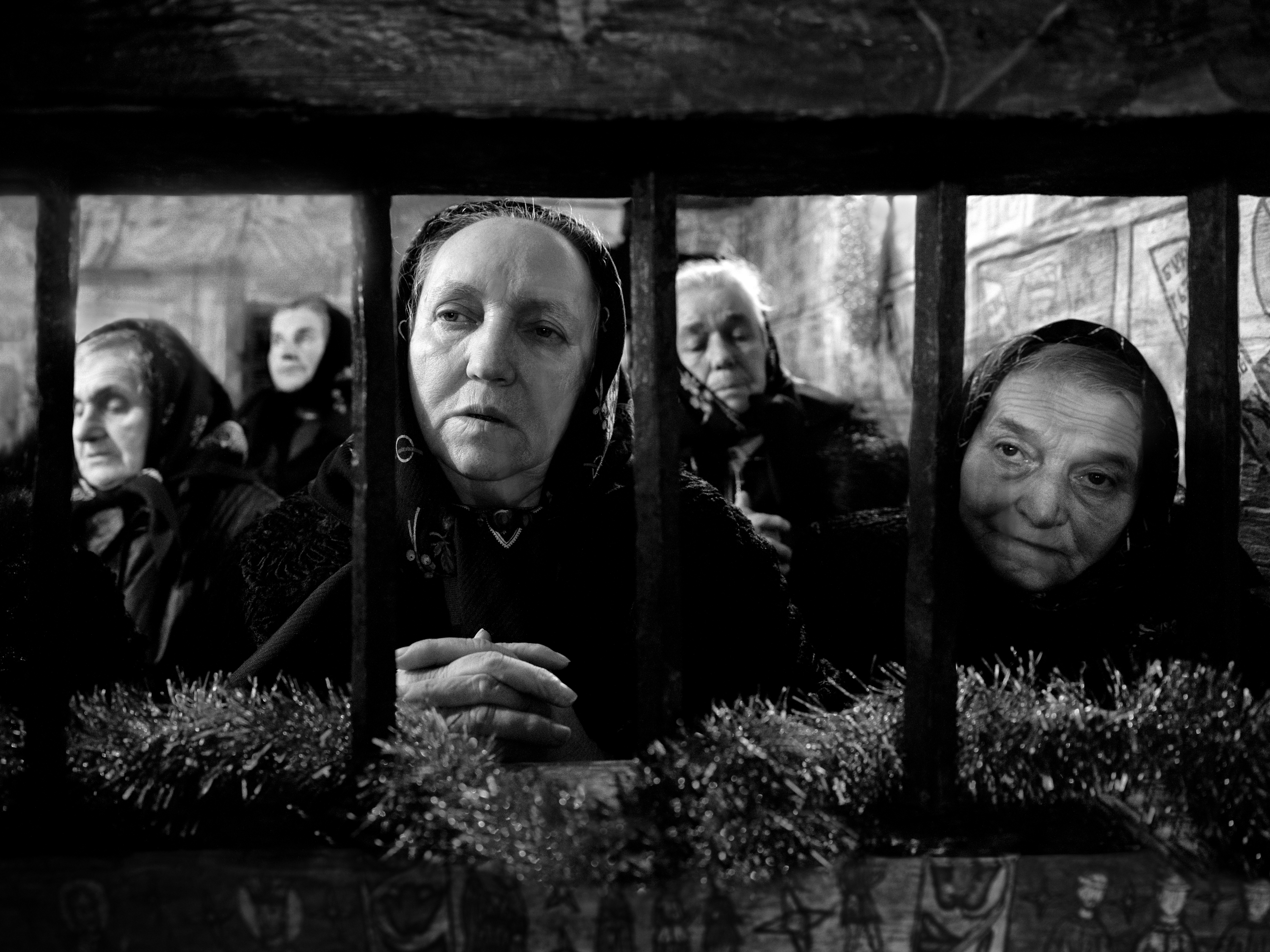 Black and white photograph of a row of elderly women in headscarves pressing close to an iron church gate strung with tinsel, their faces lit against the dark interior.
