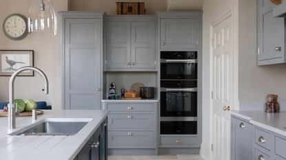 A general interior view of a domestic dove grey kitchen, with painted white walls, island with quartz worktop, chrome tap, grey crushed velvet bar stools, stainless steel range cooker oven, chrome tap, skylight, vertical radiator, potted plant and patio doors into the back garden within a home