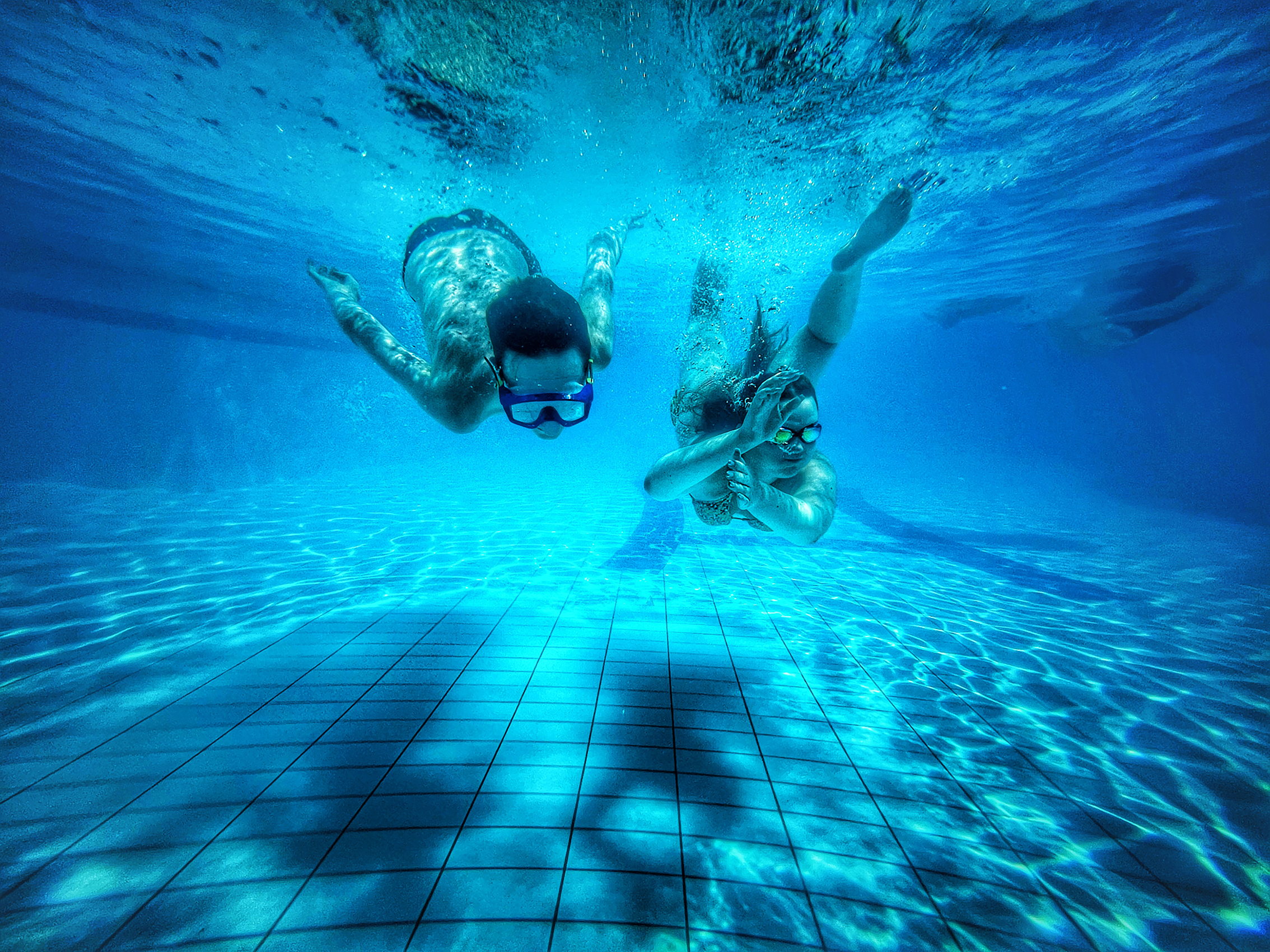 Two people wearing snorkels swim underwater in a clear blue pool, sunlight creating patterns on the tiled floor