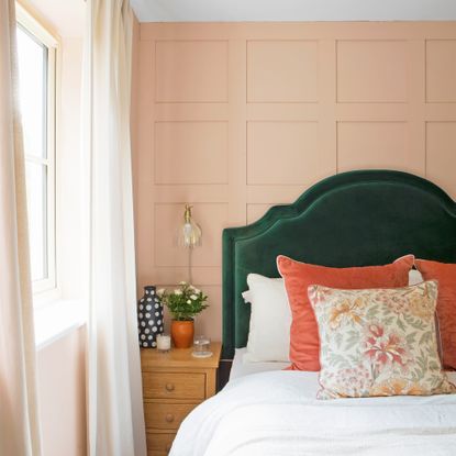 A bedroom with a panelled wall painted in peach paint, and a bed with a green upholstered headboard and orange cushions
