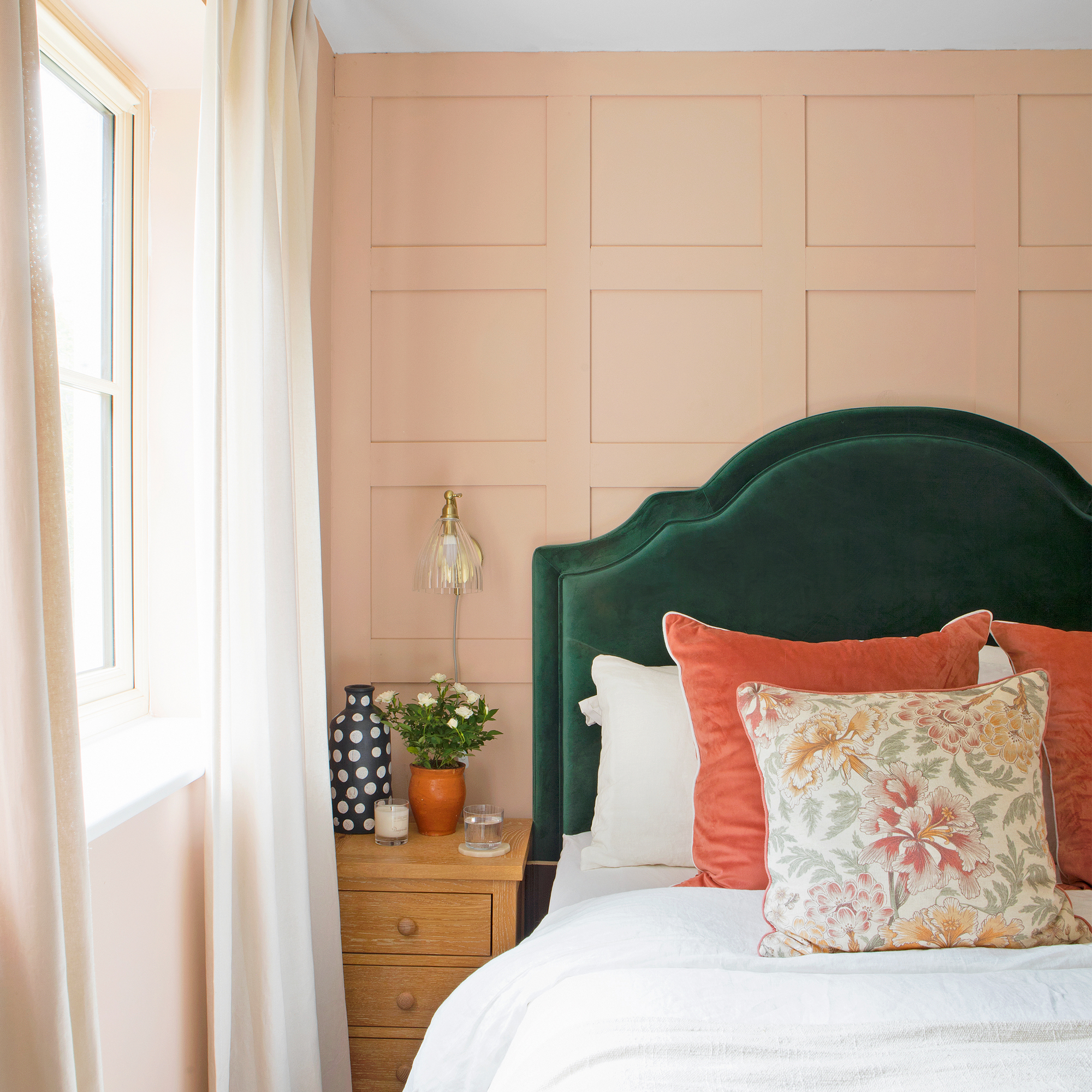 A bedroom with a panelled wall painted in peach paint, and a bed with a green upholstered headboard and orange cushions