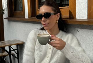 Woman wearing white sezane cardigan and black sunglasses, holding up coffee cup.