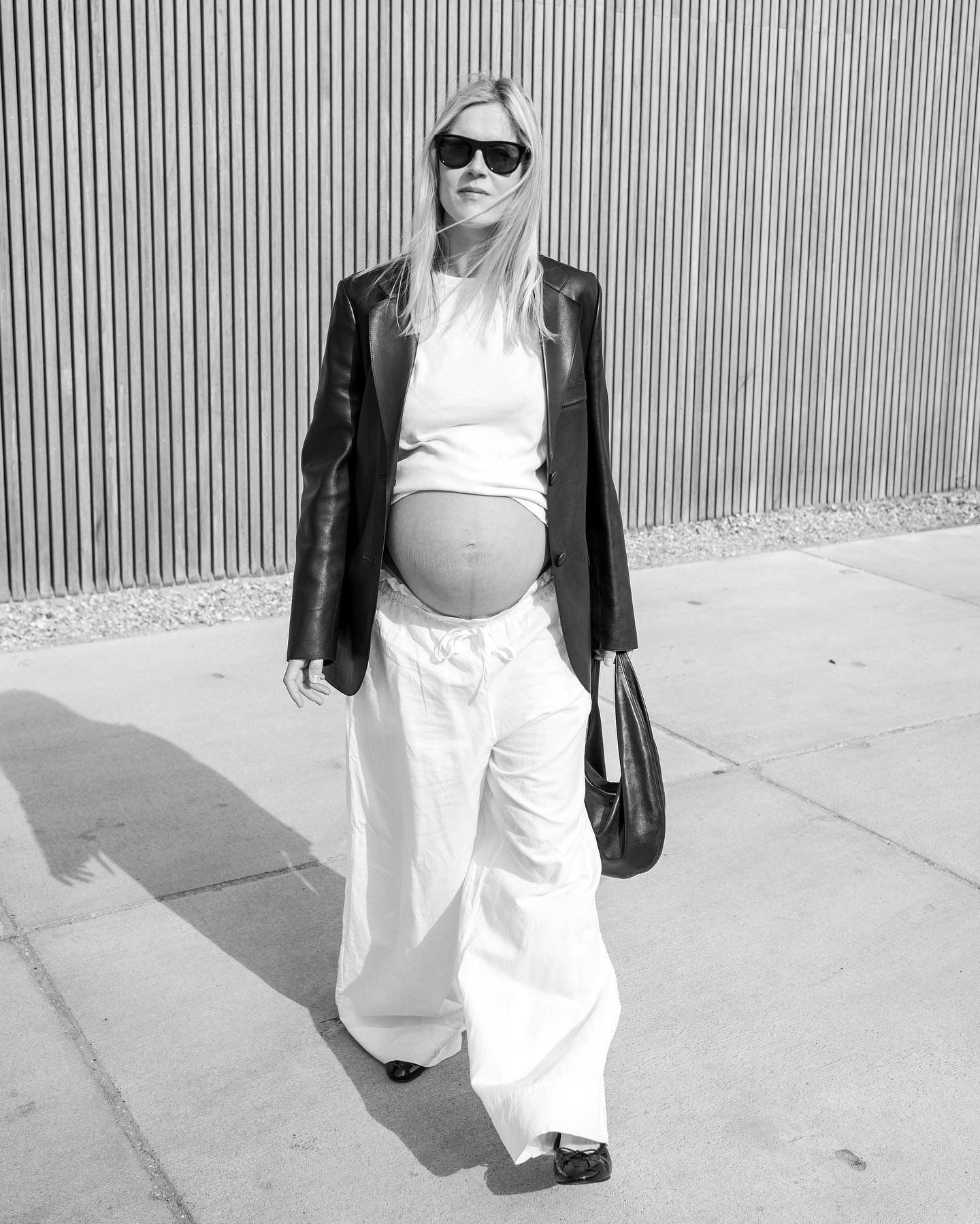 Dutch style influencer Linda Tol poses in Amsterdam wearing a pregnancy outfit with black sunglasses, a black leather blazer, cropped white T-shirt, a black leather shoulder bag, relaxed white drawstring pants, and black ballet flats