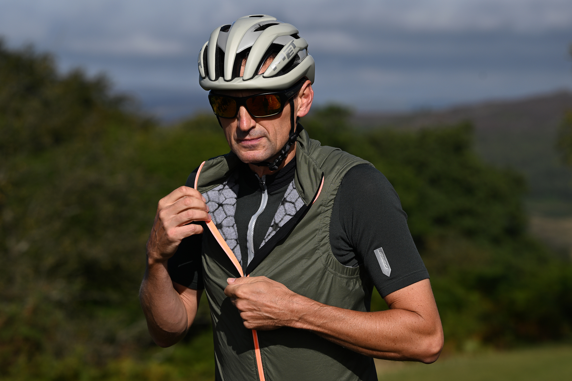 Man unzipping a green gilet and wearing dark green jersey, beige helmet and sunglasses