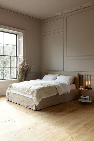 neutral bedroom with wooden floor, stone coloured bed with white and neutral bedding, side table with lamp. Stone coloured walls