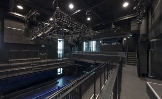 Interior image of the Cultural Centre, Dordrecht, dark room, dark grey walls, ceiling and floor, viewing gallery and safety barrier, metal ceiling framework with stage lighting, ceiling spotlights, looking down to the ground floor with blue lighting, steps, windows