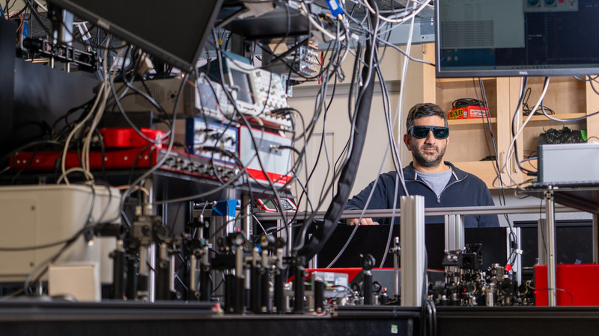 A man wearing dark safety glasses stands behind a table full of wires, mirrors, and lasers