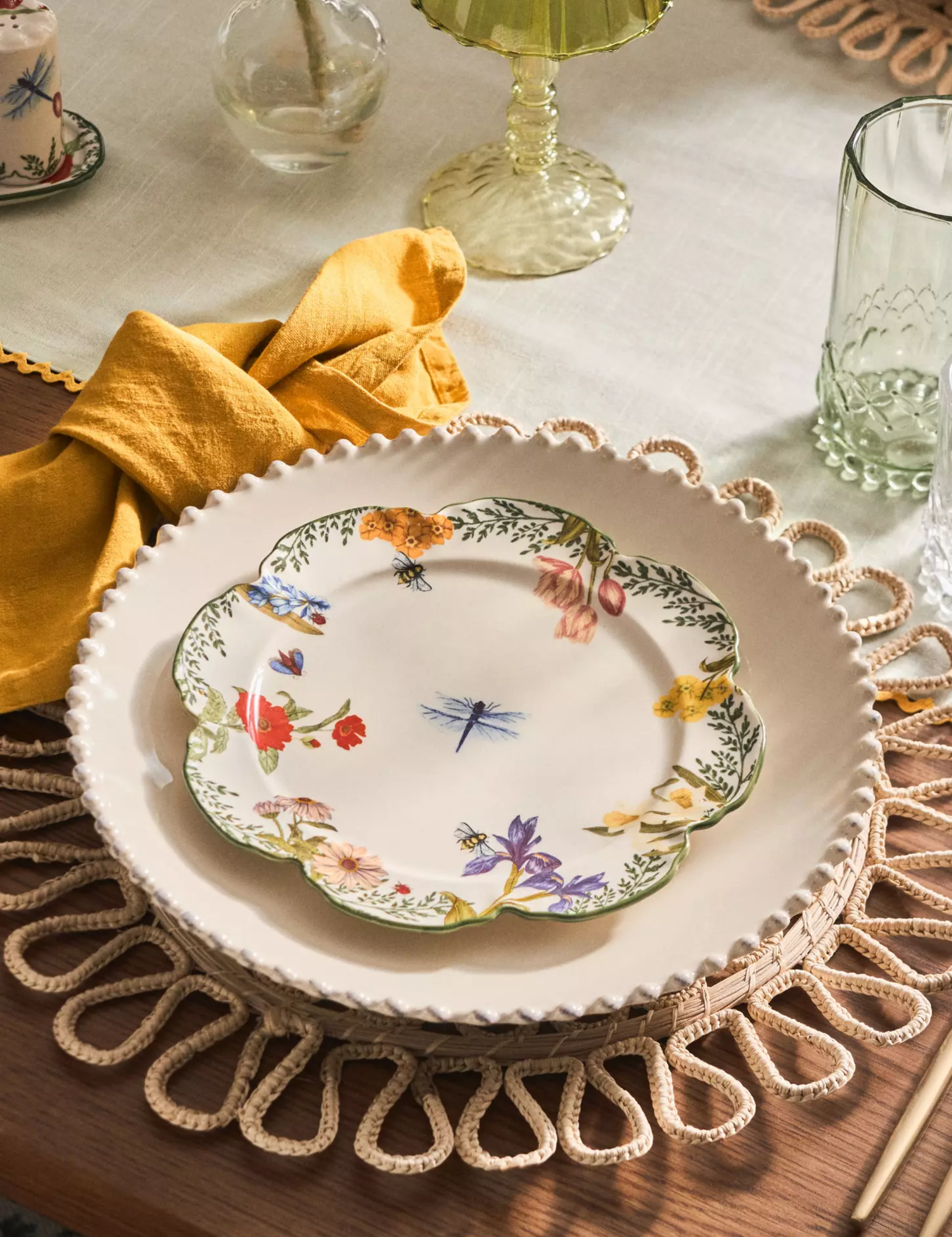 Eclectic table setting featuring a scallop-edge woven charger, floral plates, yellow linen napkins, and crystal-cut glassware