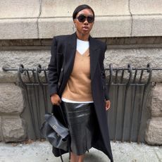 Woman wears black coat, white T-shirt, camel jumper, black skirt and black handbag