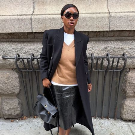 Woman wears black coat, white T-shirt, camel jumper, black skirt and black handbag
