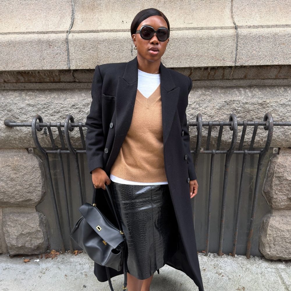Woman wears black coat, white T-shirt, camel jumper, black skirt and black handbag