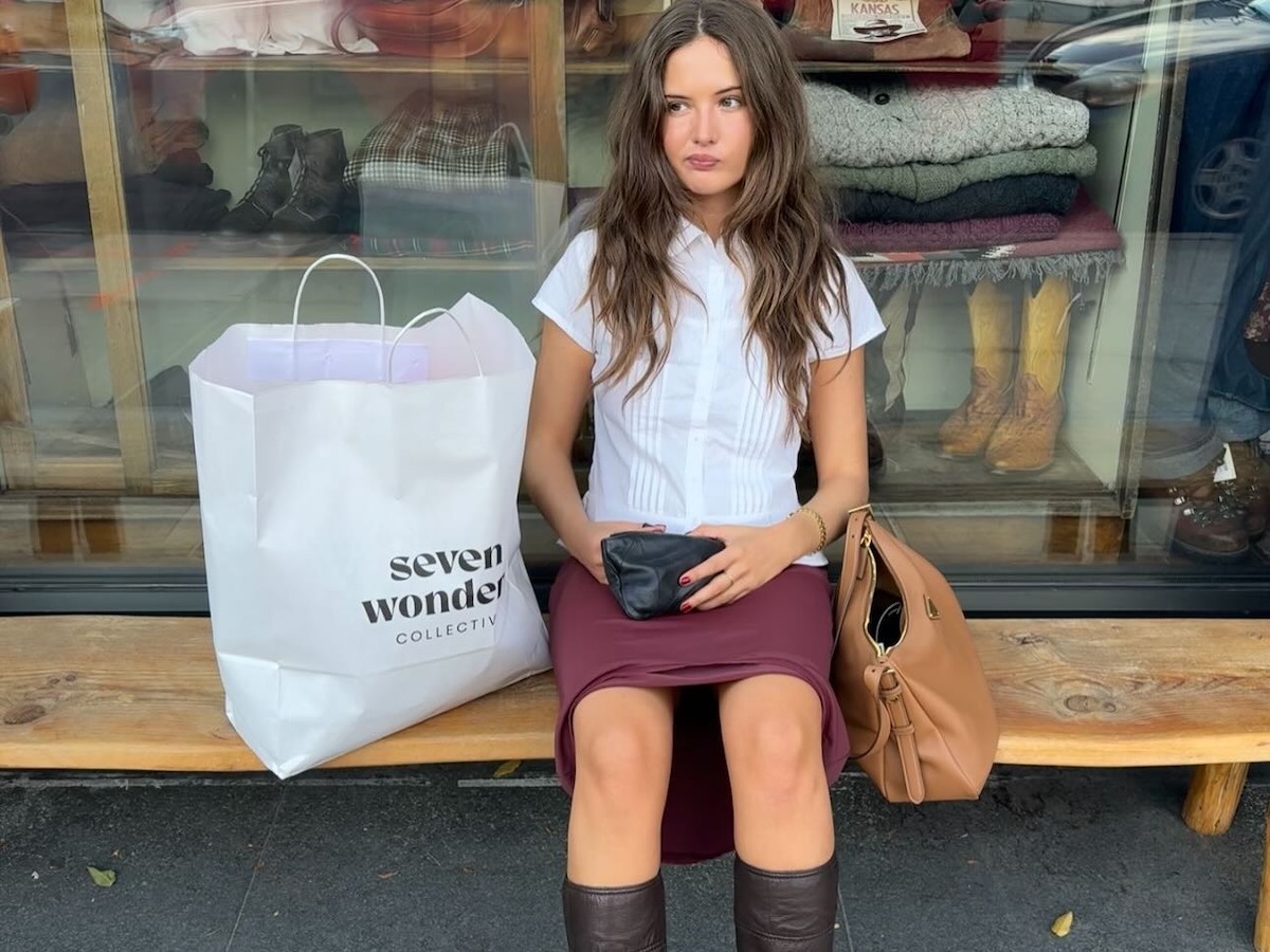 A woman sitting on a bench wearing a white button-up blouse, a red skirt, and brown knee-high boots.