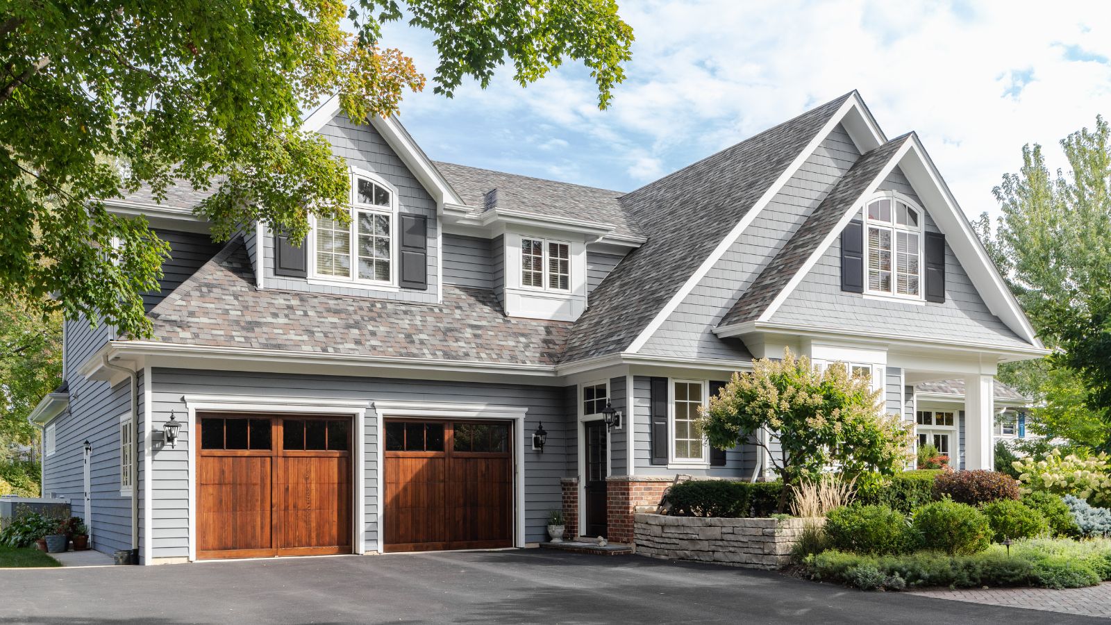 A large grey and white modern farmhouse with two wooden garage doors, and beautiful landscaping.