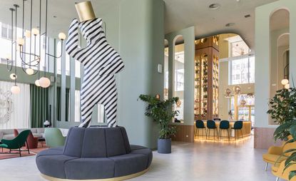 A hotel sitting area with a range of different size and color chairs, a round grey sofa with a striped human shaped art model on it, a bar, potted plants and artistic round pendant lights.