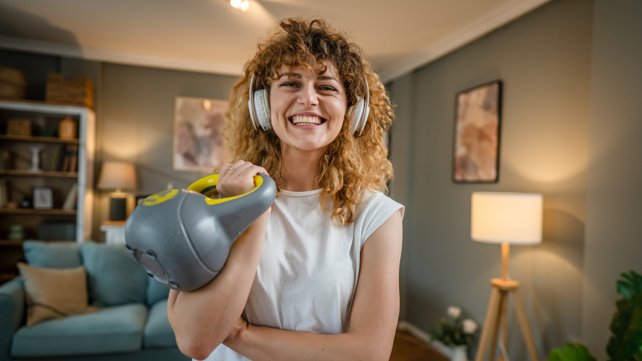 Woman wearing headphones and smiling at viewer holds kettlebell by her shoulder. She is in a domestic setting.