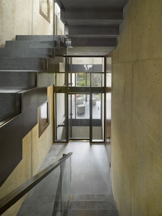 Left of the image is a grey stair case going up and on the right is a grey staircase going down towards the building exit. The staircase have glass banisters. Exit doors are floor to ceiling glass doors