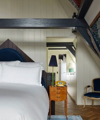 neutral bedroom with panelled walls and hints of blue in the headboard and upholstery with fresh white bed linen and a view through a doorway into a storage wardrobe area