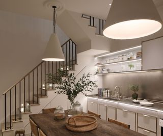 A cosy kitchen-dining area featuring a rustic wooden table with woven chairs, adorned with glass jars and a woven tray. Above, two large conical pendant lights provide warm illumination. The background includes a staircase with black railings and a modern kitchen with open shelving, under-cabinet lighting, and a sleek stainless steel backsplash.