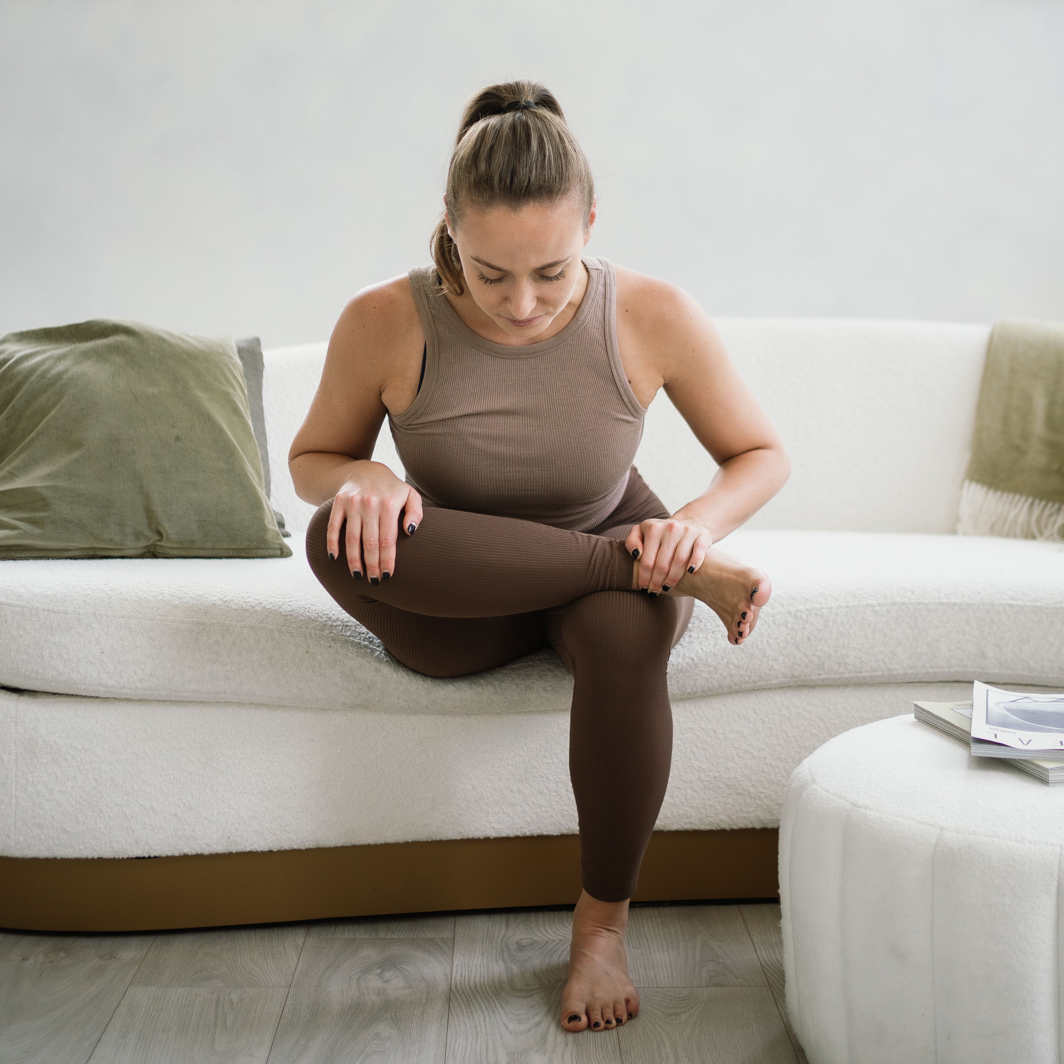 A woman sits on the edge of a couch performing a figure four fold. Her left leg is bent, foot flat on the floor, on her right ankle is crossed over her left thigh. She is holding her right knee and ankle as she leans forwards from her hips.