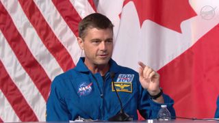 A man with gray hair wearing a blue flight suit with a circular NASA logo sits in front of the American and Canadian flags with a microphone in front of him