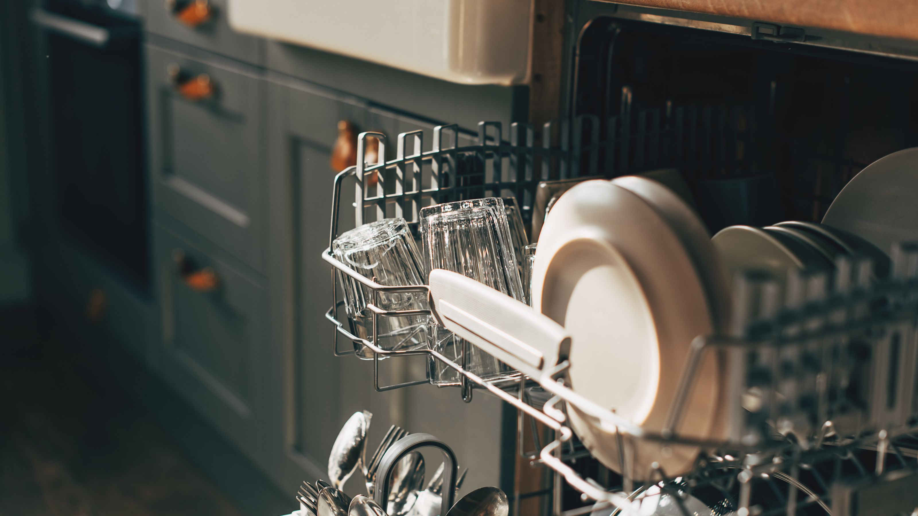 Open dishwasher with loaded top drawer pulled out in a kitchen with grey blue cupboards and a butlers sink