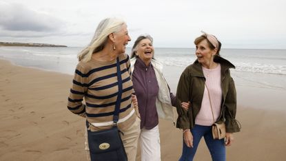 Three older women laugh as they walk along the beach.