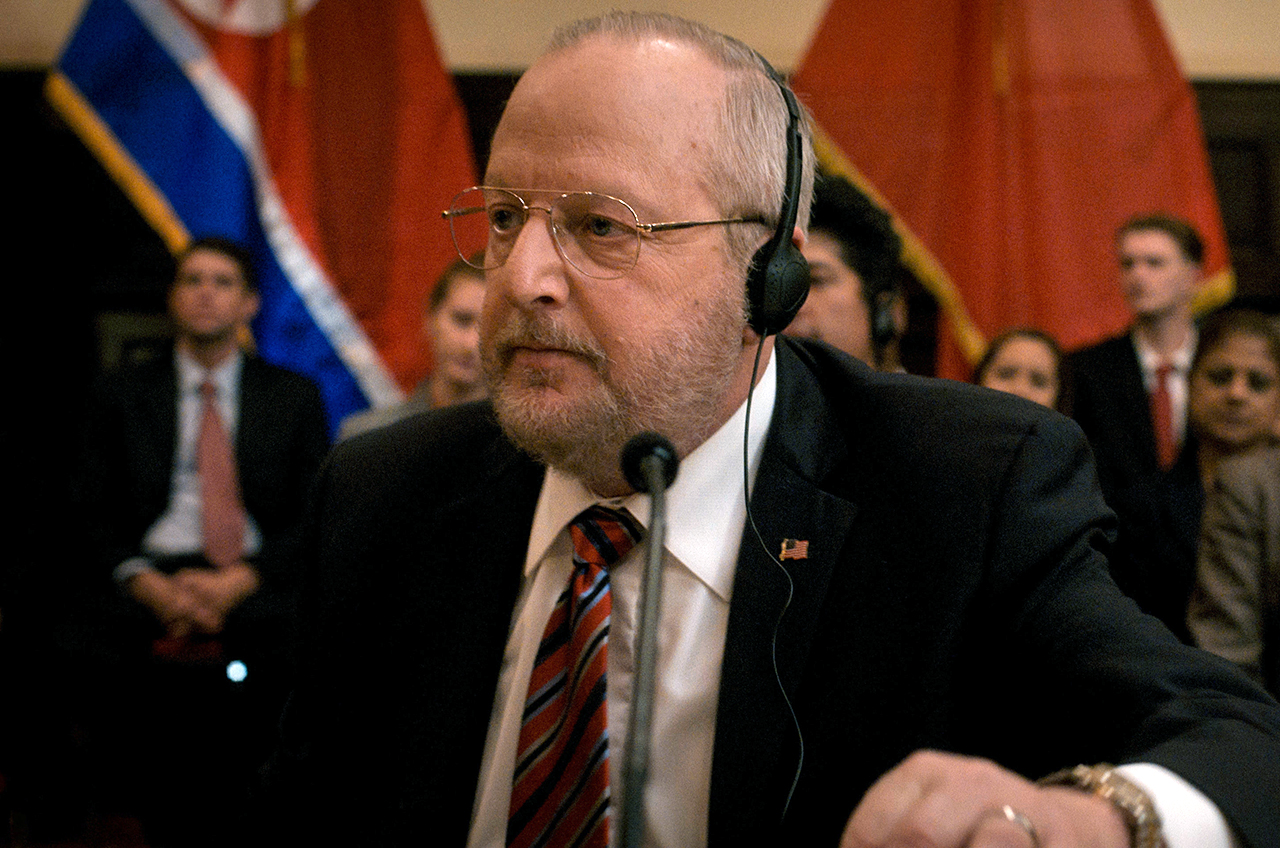 photo of an older man in a suit sitting down and wearing dark headphones.
